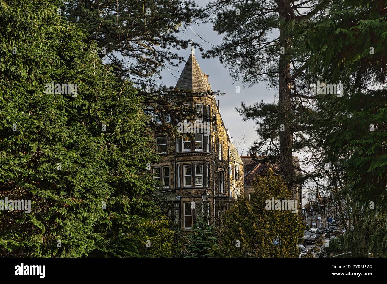 Un bâtiment en pierre avec un toit conique proéminent est partiellement visible à travers les branches des arbres à feuilles persistantes. Le bâtiment dispose de plusieurs fenêtres et Banque D'Images