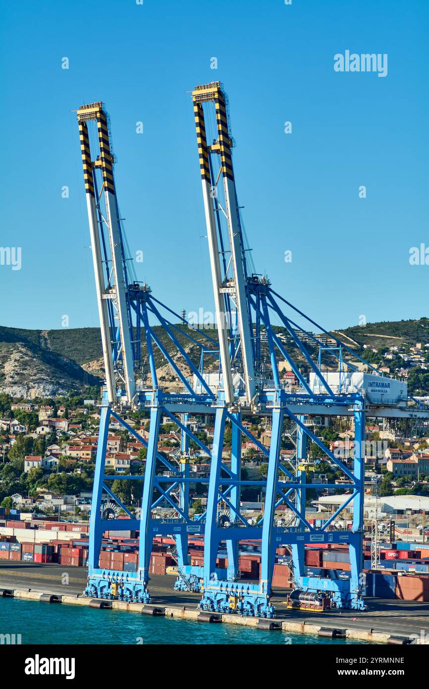 Marseille, France, 4 décembre 2024 : deux portiques bleus à bandes noires-jaunes fonctionnent dans un port très fréquenté. Les collines résidentielles forment la toile de fond, se mélangeant Banque D'Images