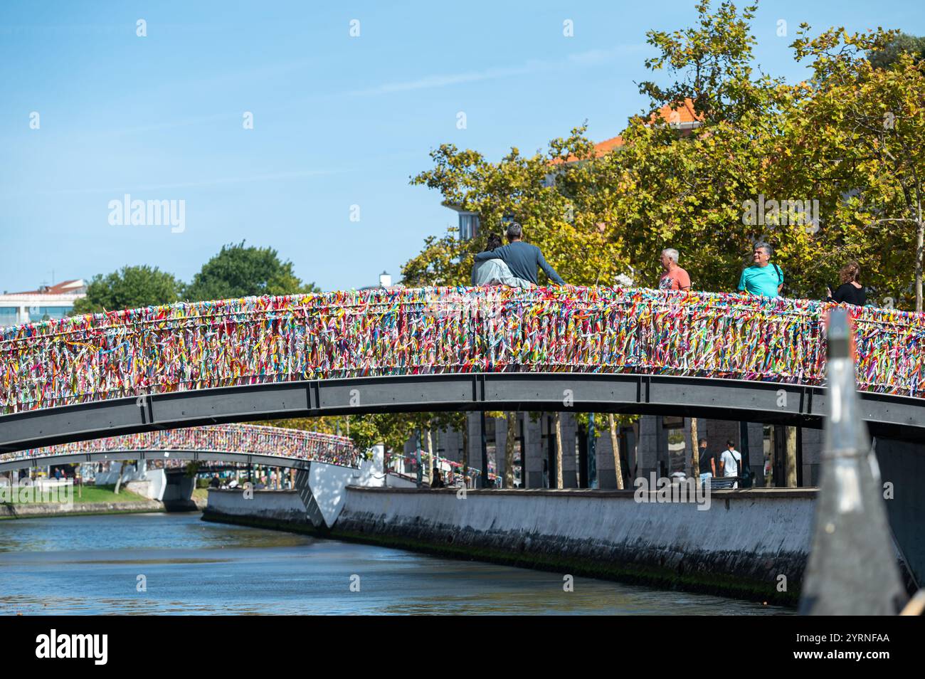 Aveiro, Portugal - 11 septembre 2024 : les couples marchent sur un pont orné d'écluses d'amour vibrantes à côté d'un paisible canal. Banque D'Images
