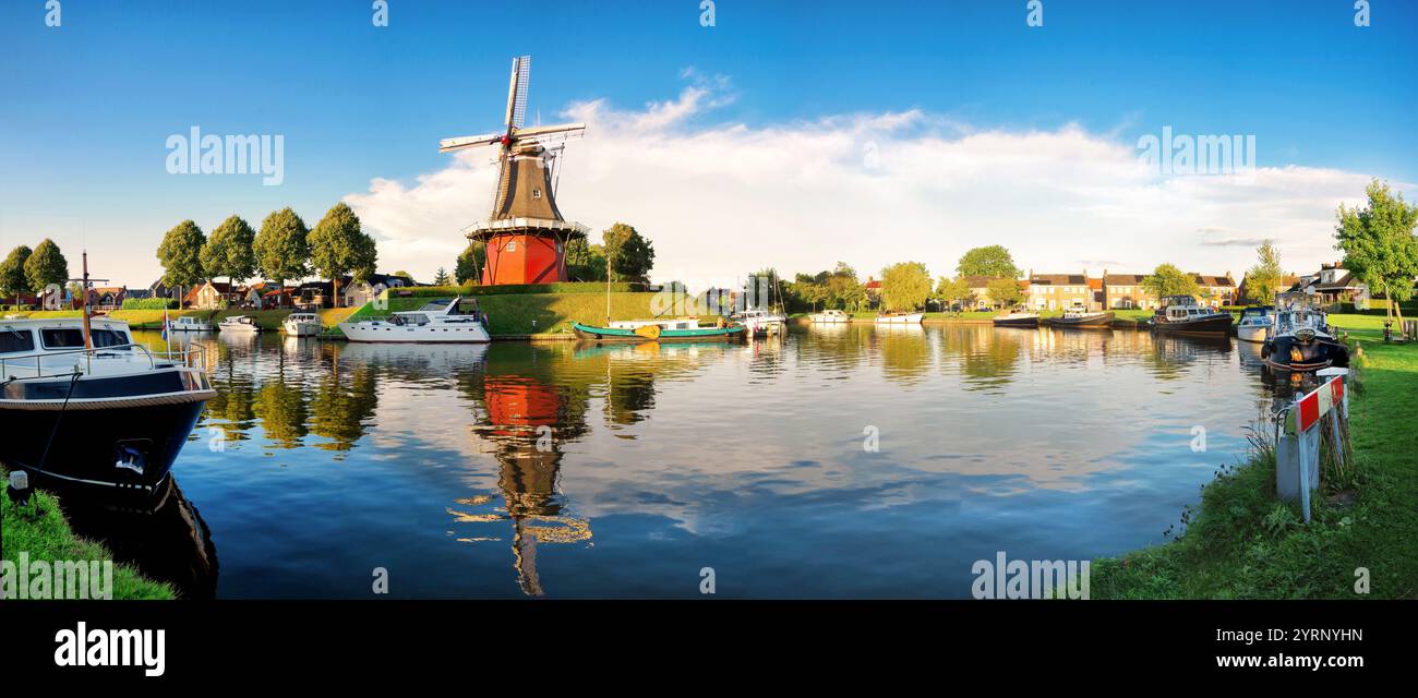 Panorama d'une voie navigable hollandaise paisible flanquée de moulins à vent et de bateaux sous un ciel dégagé à Dokkum, Frise, pays-Bas Banque D'Images
