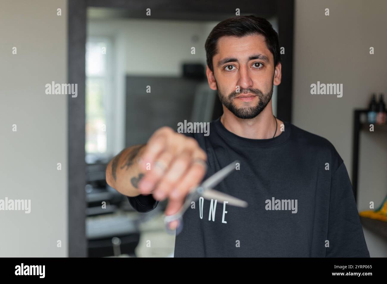 Beau gars de salon de coiffure en T-shirt noir tient des ciseaux et regarde la caméra. Banque D'Images