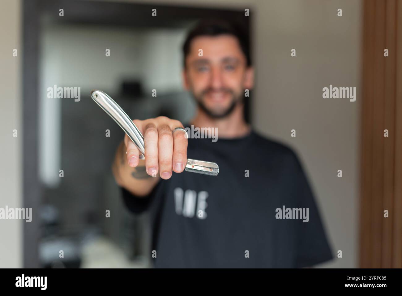 Heureux homme de barbier avec un sourire tient une lame de rasoir devant la caméra de l'objectif dans le salon de coiffure Banque D'Images