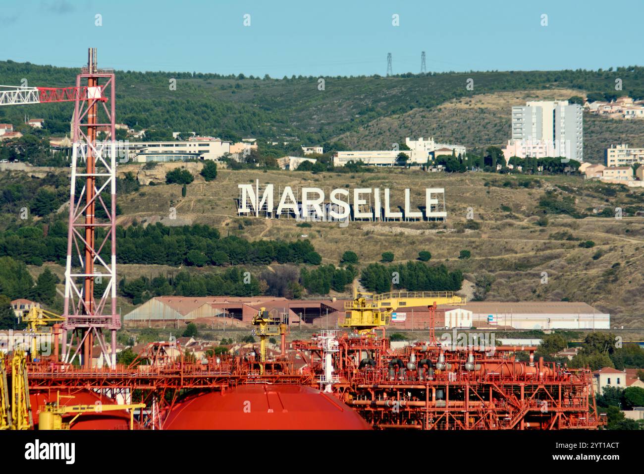 Le panneau Marseille se distingue sur une colline verdoyante, donnant sur une zone industrielle avec grues et réservoirs de stockage, avec des bâtiments résidentiels visibles dedans Banque D'Images