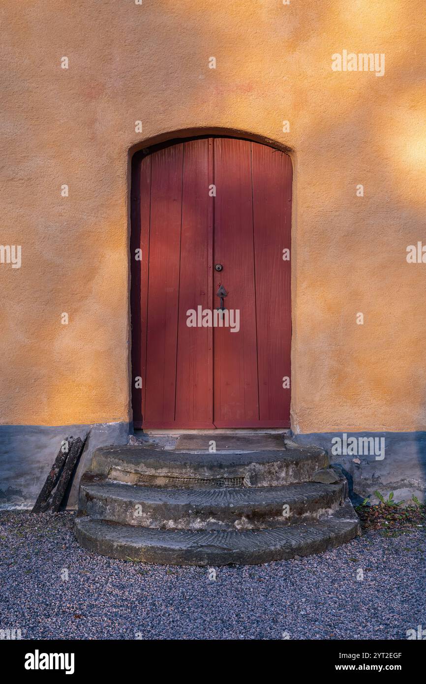 Une porte rustique en bois rouge avec un sommet cintré, située dans un mur de plâtre jaune, entourée de marches en pierre Banque D'Images