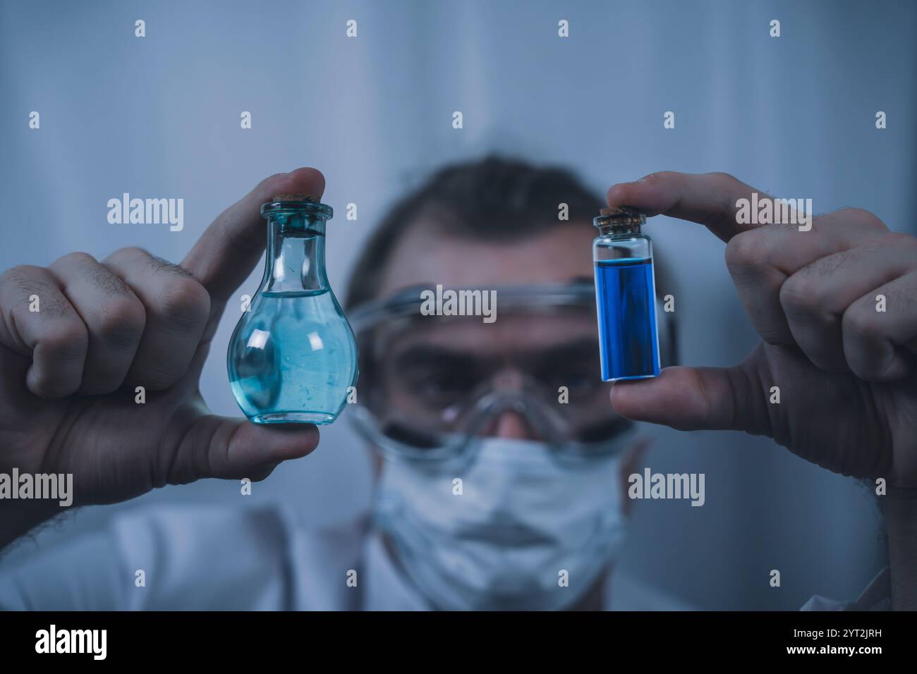 Un scientifique portant des lunettes de protection et un masque facial examinant de près deux flacons contenant des liquides bleus et clairs, mettant en évidence la recherche, la chimie Banque D'Images