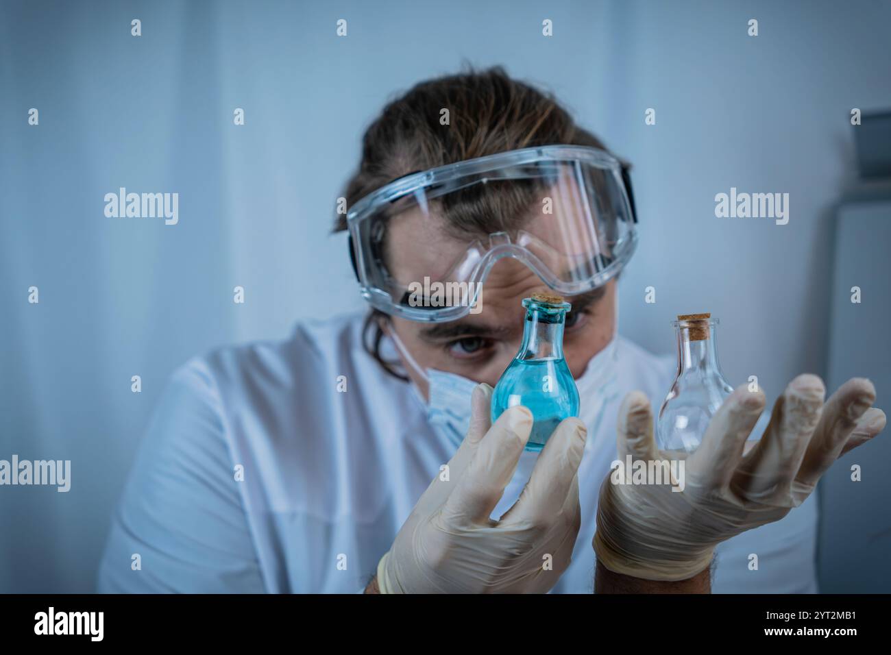Un scientifique portant des lunettes de protection, des gants et un masque facial analysant soigneusement les flacons contenant des liquides bleus et clairs, en mettant l'accent sur la précision, re Banque D'Images