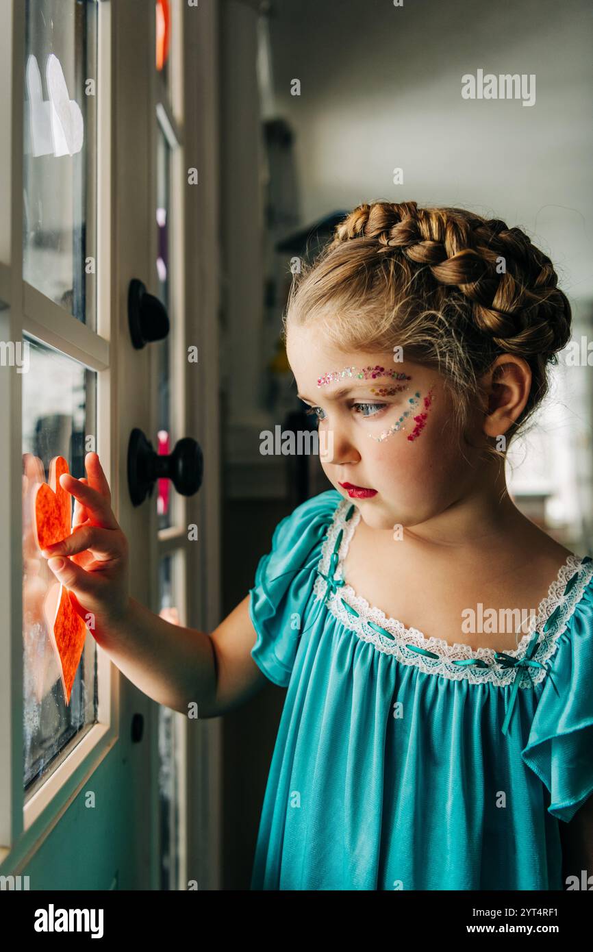 Fille avec les cheveux tressés et le maquillage de paillettes touche le coeur sur la fenêtre Banque D'Images