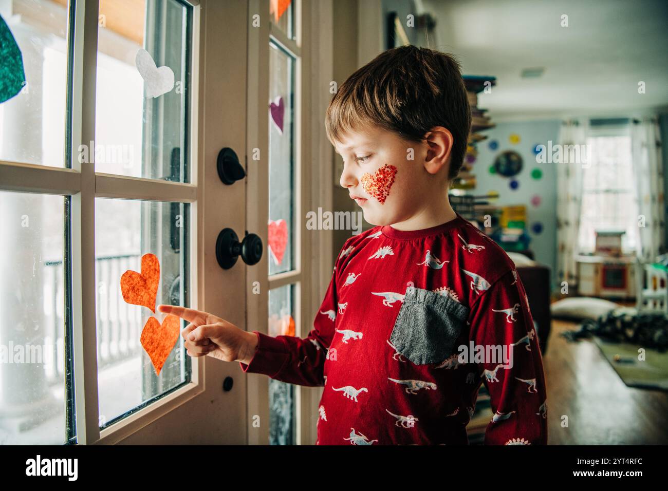 Garçon en chemise de dinosaure rouge avec coeur pailleté touche les coeurs de fenêtre Banque D'Images