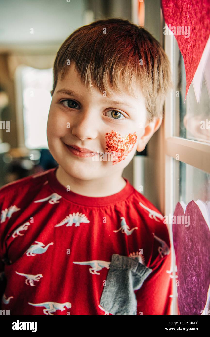 Garçon souriant en chemise de dinosaure rouge avec maquillage coeur pailleté par fenêtre Banque D'Images