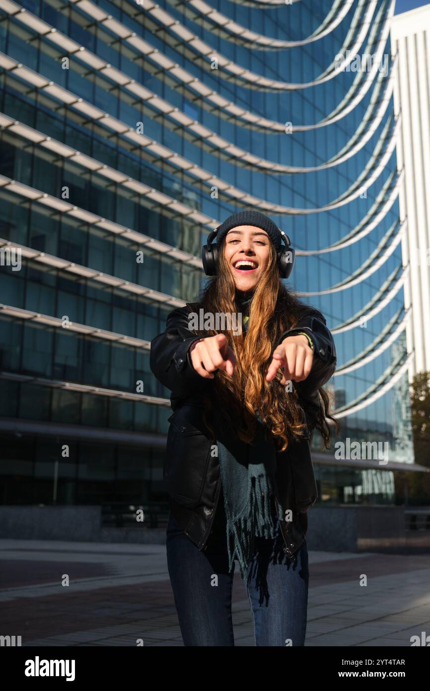 Femme porter des écouteurs pointe vers vous et sourire devant un bâtiment Banque D'Images
