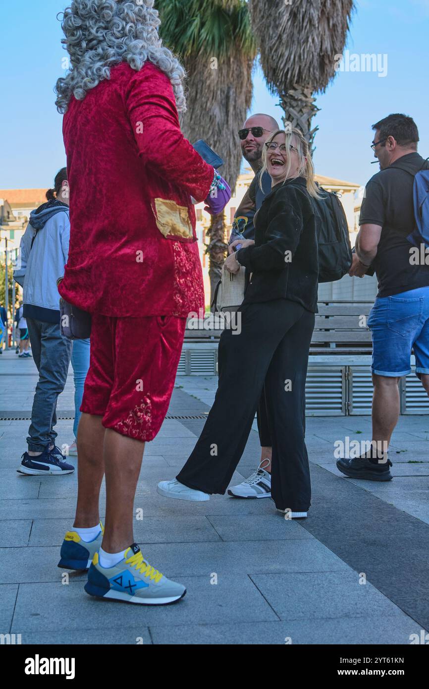 Sabona. Italia - 06 décembre 2024 : un individu vêtu d'un costume de velours rouge théâtral avec des baskets modernes sur une place publique, entouré de c. Banque D'Images
