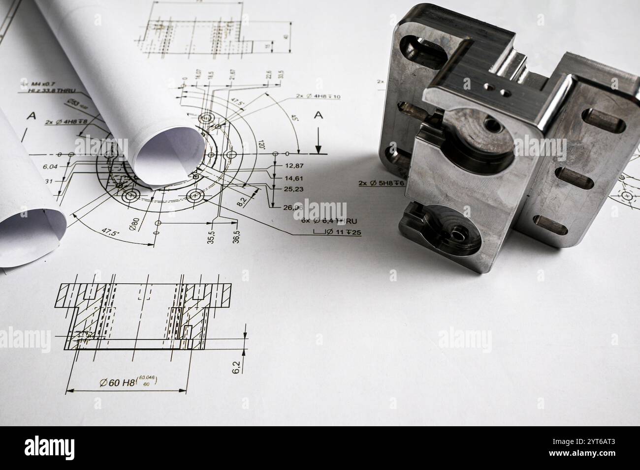 Pièce métallique reposant sur un dessin d'ingénierie avec des plans roulés, illustrant l'ingénierie mécanique et la fabrication de précision. Banque D'Images