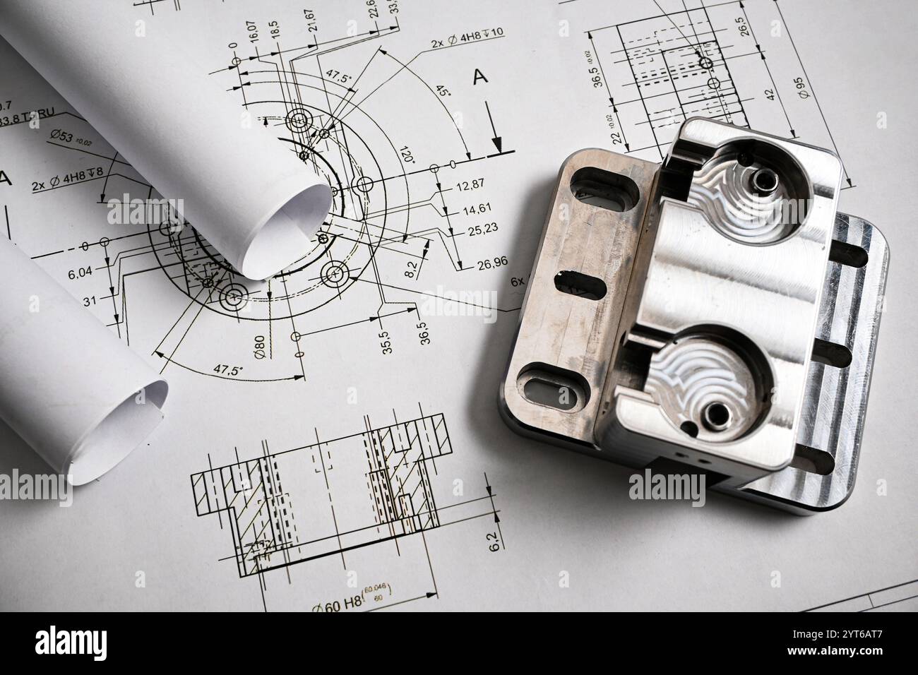Pièce métallique reposant sur un dessin d'ingénierie avec des plans roulés, illustrant l'ingénierie mécanique et la fabrication de précision. Banque D'Images