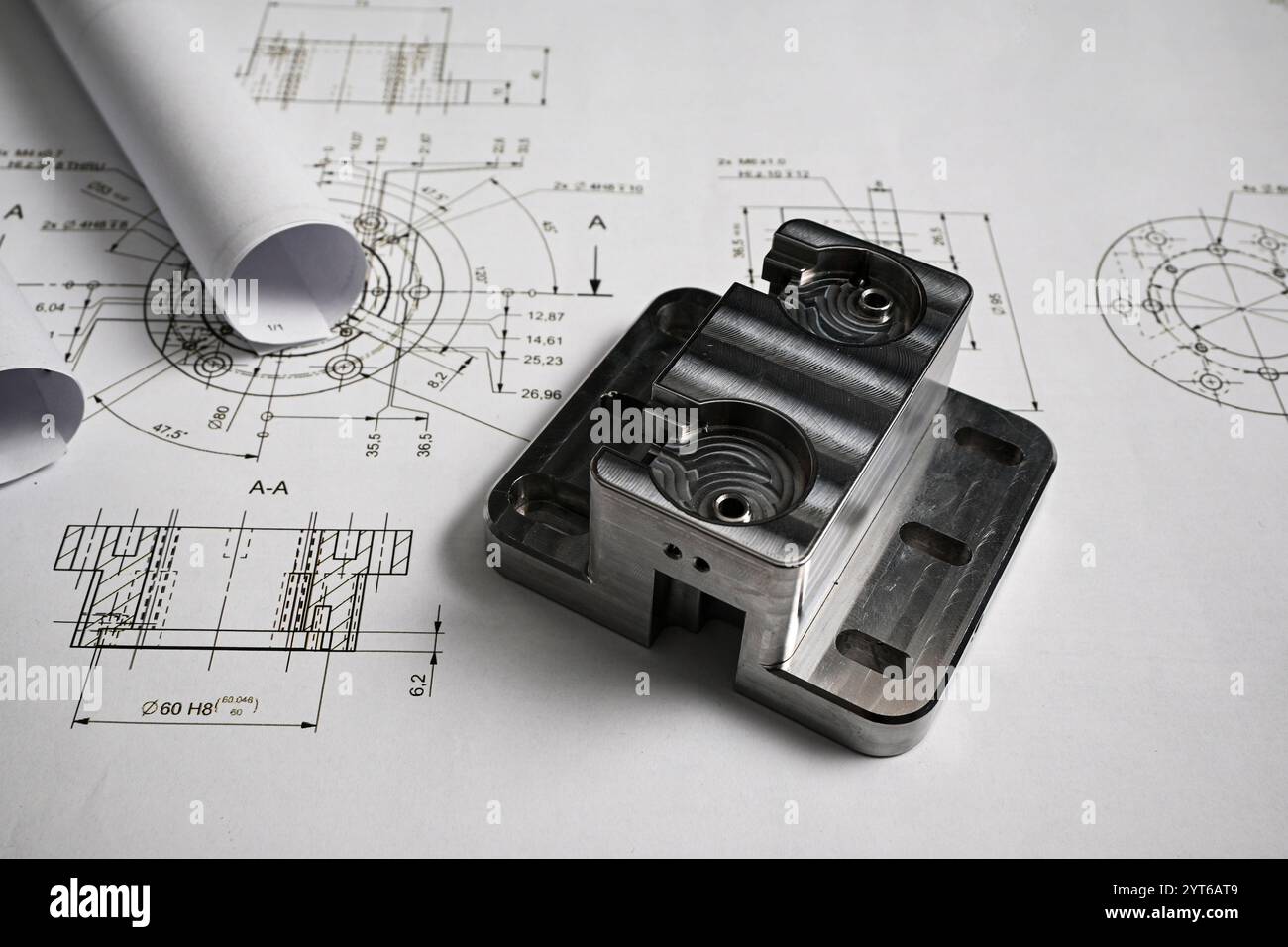 Pièce métallique reposant sur un dessin d'ingénierie avec des plans roulés, illustrant l'ingénierie mécanique et la fabrication de précision. Banque D'Images