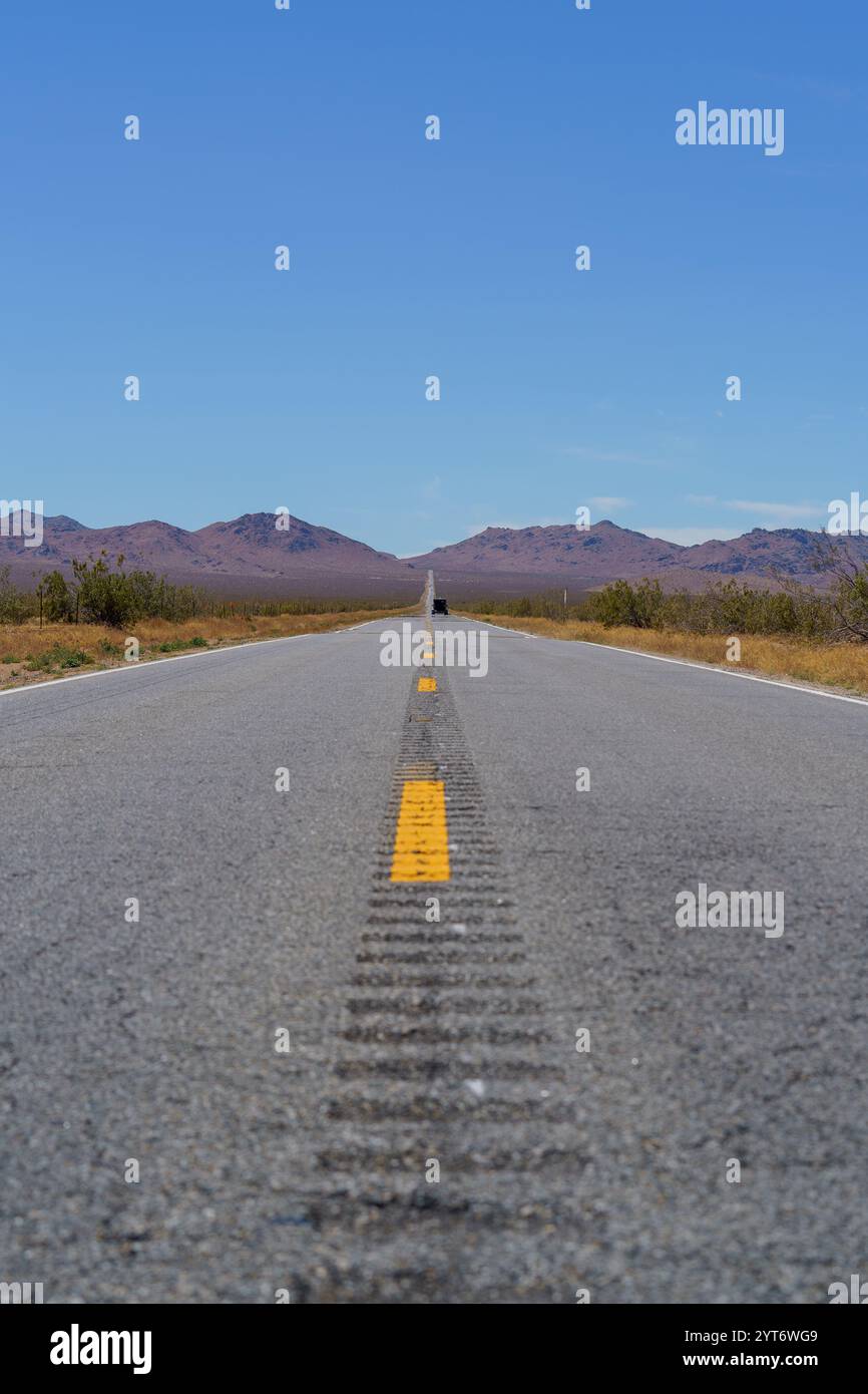 Une longue route asphaltée droite avec des lignes jaunes s'étend vers des montagnes lointaines et basses sous un ciel bleu clair dans le désert. Banque D'Images