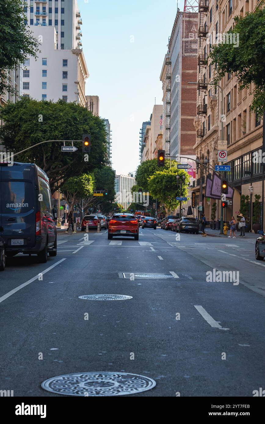 Une rue animée du centre-ville de Los Angeles dispose de grands bâtiments, d'un fourgon Amazon, de voitures, de feux de circulation et de piétons, avec des arbres bordant les trottoirs. Banque D'Images