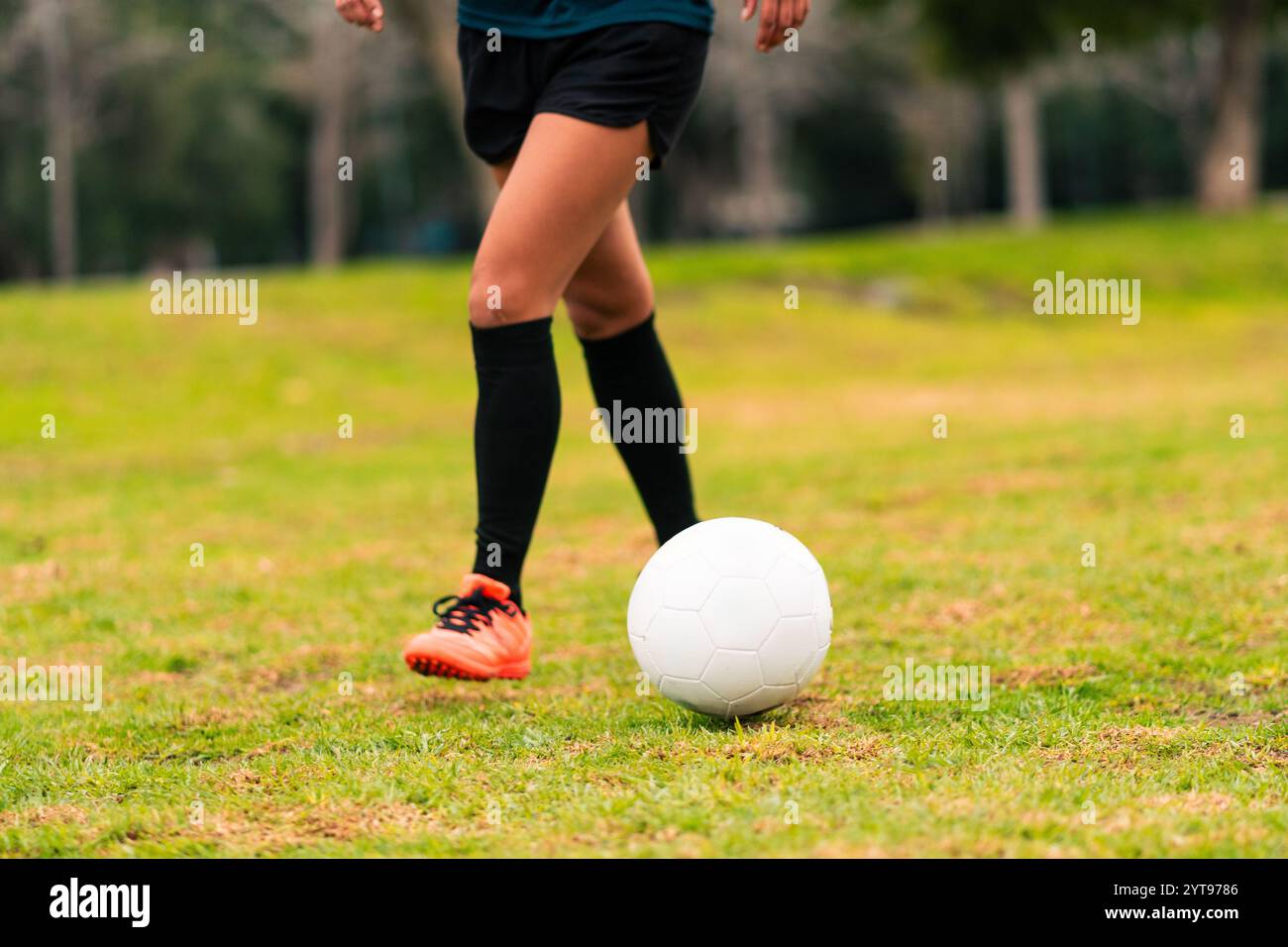 Vue des jambes d'une joueuse de football brune qui court pour donner un coup de pied au ballon de football. sur une herbe verte à l'extérieur, avec un fond coloré. avec des vêtements de sport. Banque D'Images