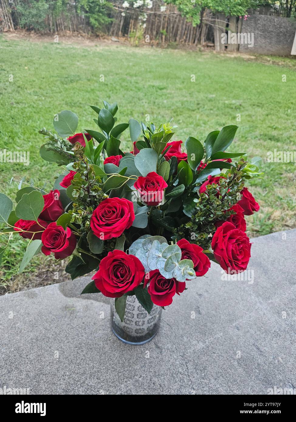 Un vase avec des roses rouges et un feuillage vert sur une table, placé sur un fond de pelouse verte et d'arbres, pris à la lumière du jour. - Image de stock capturée avec un smartphone