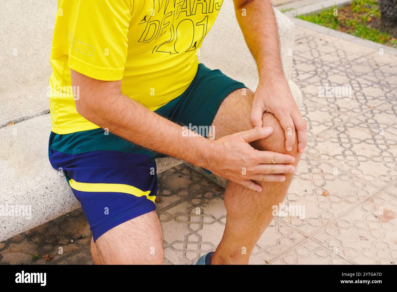 Un homme d'âge moyen qui fait du jogging se plaint de douleurs au genou et tient son genou sur un banc Banque D'Images