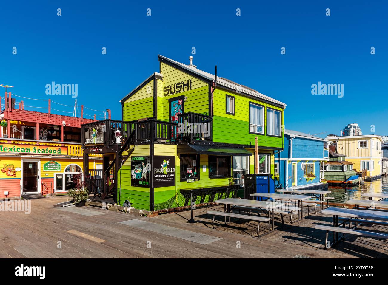 Un bâtiment coloré avec un extérieur vert et jaune et un panneau vert qui dit sushi. Le bâtiment est entouré de tables et de chaises, et il y a un Banque D'Images