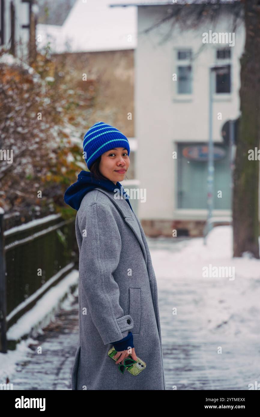 Une femme se tient debout dans une rue enneigée, portant un manteau gris et un chapeau coloré, capturant un moment serein de l'hiver. Banque D'Images