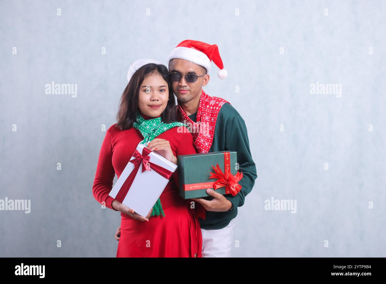 portrait de jeune couple portant des pulls de noël, des chapeaux de père noël et des foulards, souriant élégamment regardant vers l'avant portant rouge et blanc cadeau de noël b Banque D'Images