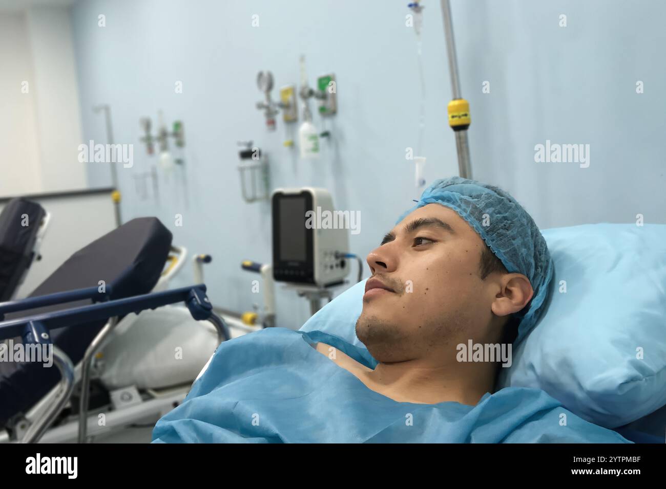 Un homme se repose sur un lit d'hôpital, portant une blouse médicale et une casquette chirurgicale. La salle est équipée de matériel médical, y compris des moniteurs et IV Banque D'Images