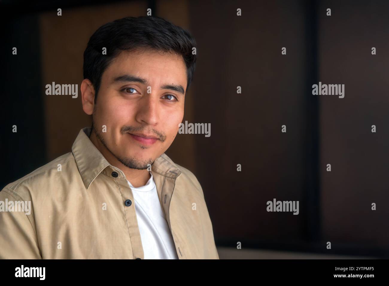 Un jeune homme se tient à l'intérieur, exsudant la confiance avec un sourire chaleureux. Il porte une chemise légère sous une veste beige, complétée par une atmosphère accueillante Banque D'Images