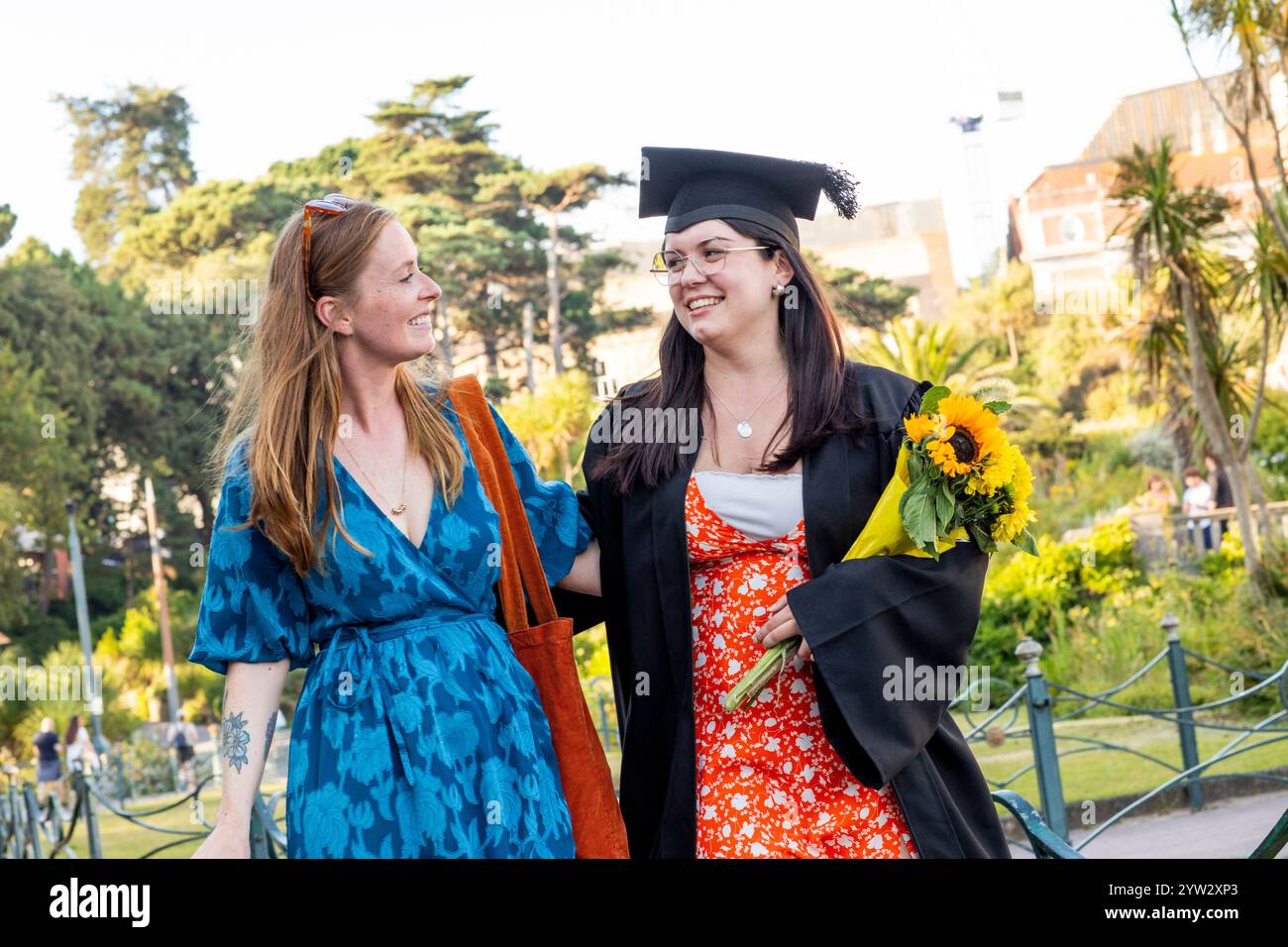 Deux femmes souriantes marchant dans un parc, une dans une casquette de remise de diplôme et une robe tenant un bouquet de tournesol, Bournemouth, Dorset UK Banque D'Images