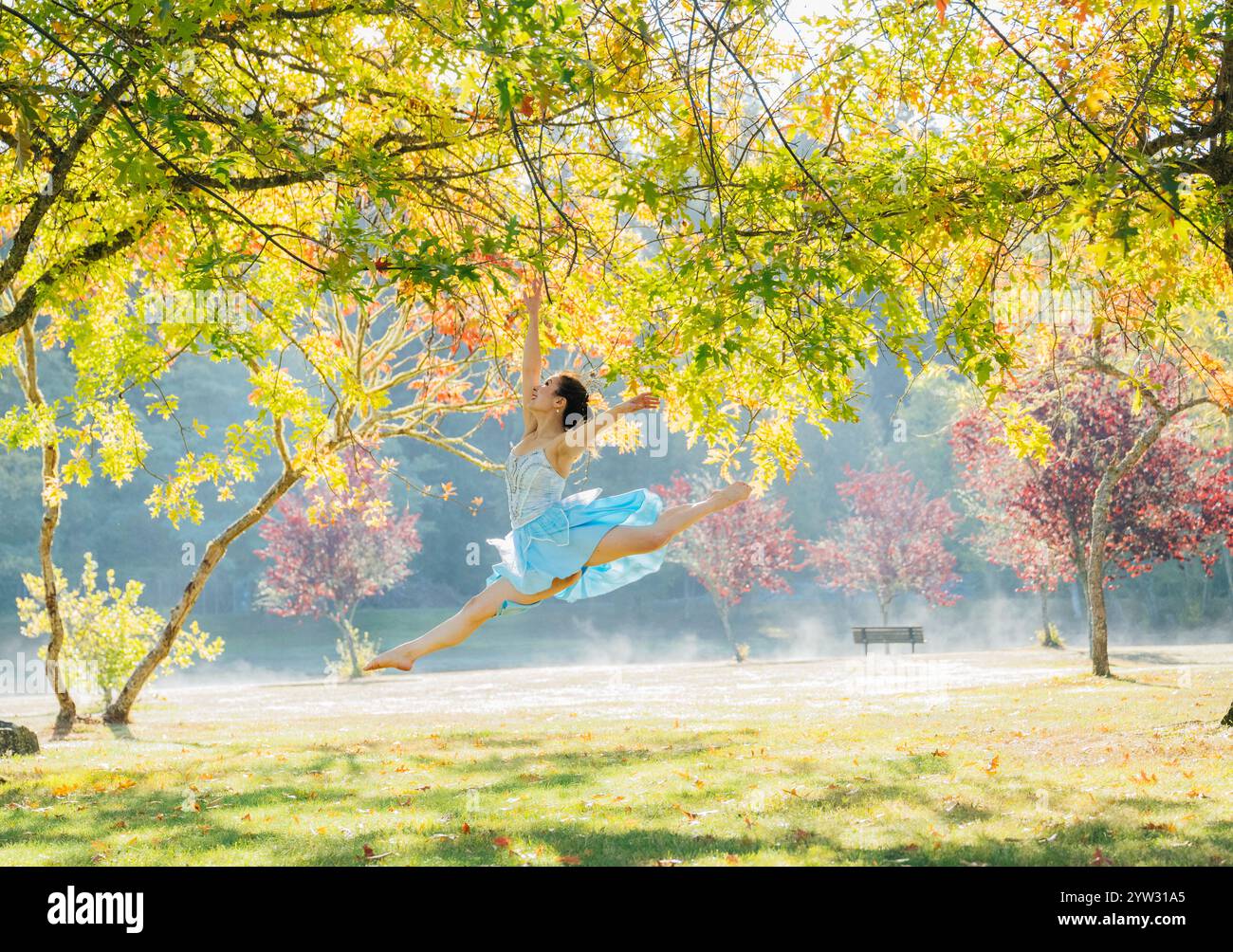 Femme dans une robe bleue sautant gracieusement dans un parc ensoleillé avec du feuillage d'automne. Banque D'Images