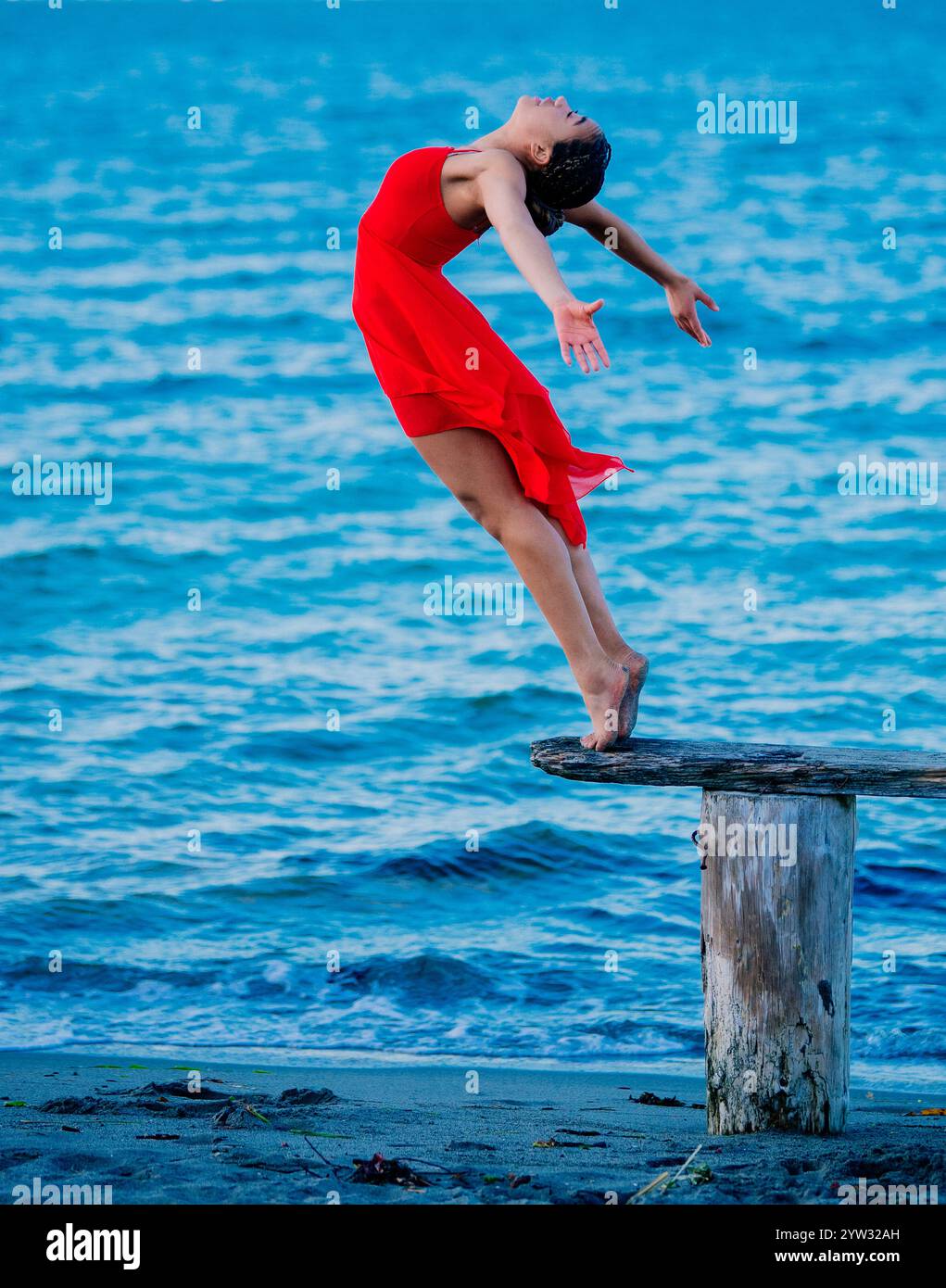 Danseuse gracieuse dans une robe rouge effectuant un mouvement de danse sur un moignon en bois par la mer bleue, USA Banque D'Images