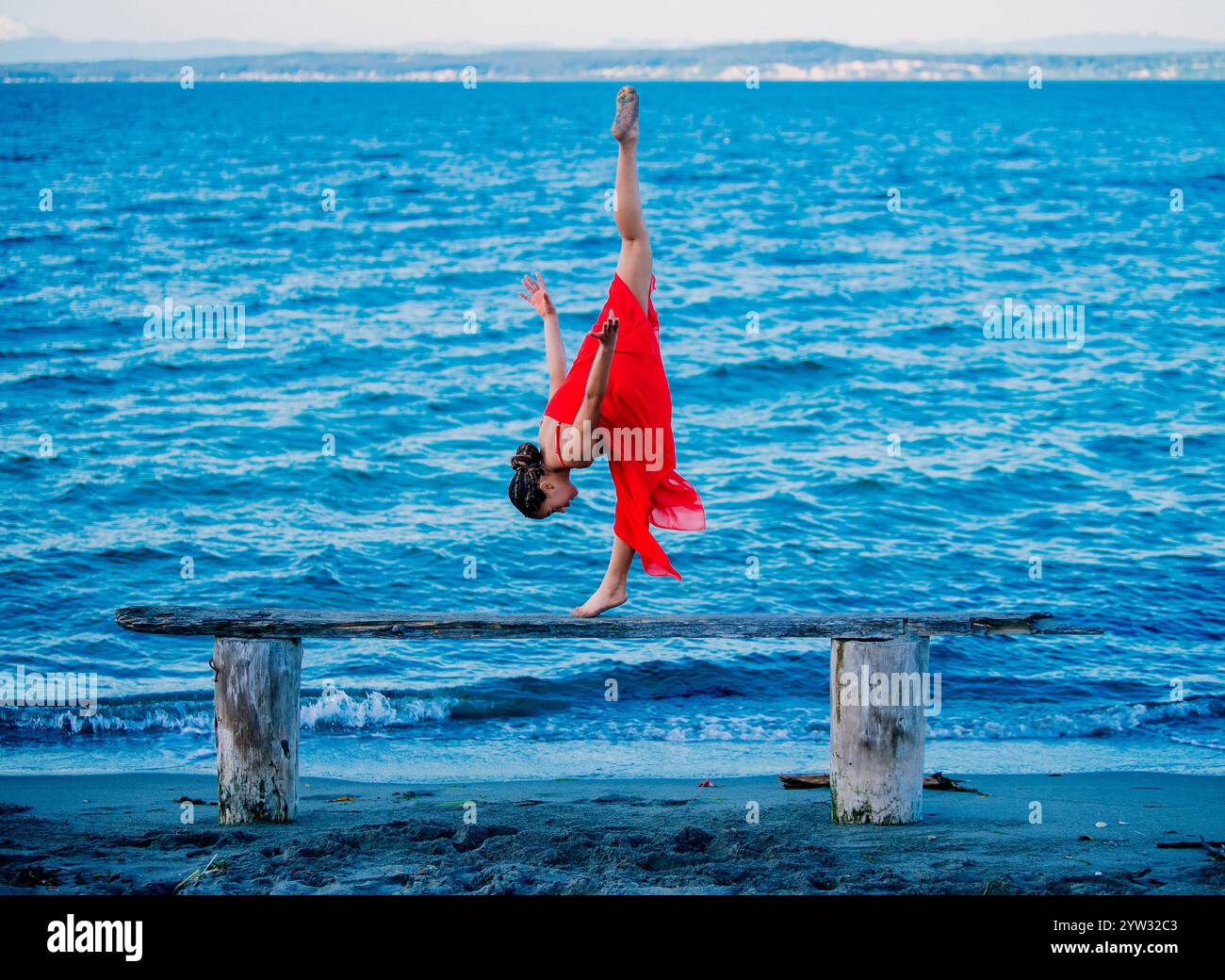 Femme dans une robe rouge exécutant un stand acrobatique sur une poutre en bois au bord de la mer, USA Banque D'Images