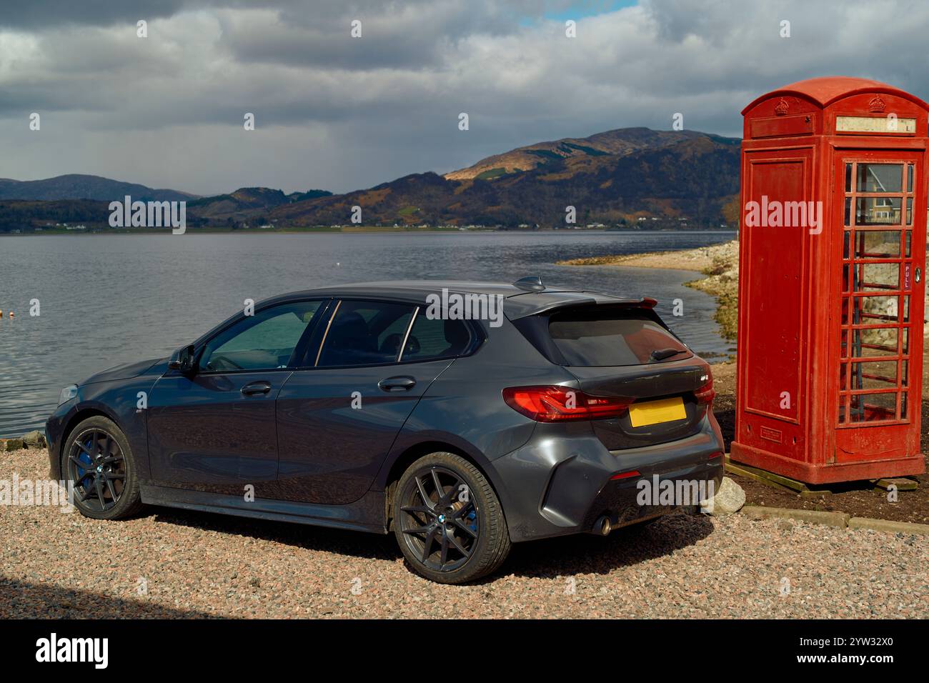 Une voiture à hayon grise moderne garée à côté d'une cabine téléphonique rouge traditionnelle avec un lac pittoresque et des montagnes en arrière-plan, Highlands, Écosse Banque D'Images