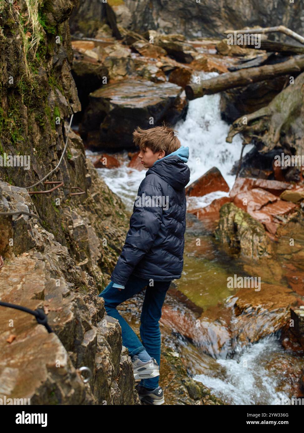 Adolescent dans une veste marine et un Jean bleu se tient sur un rocher par un ruisseau coulant avec des pierres rouges, Écosse Banque D'Images