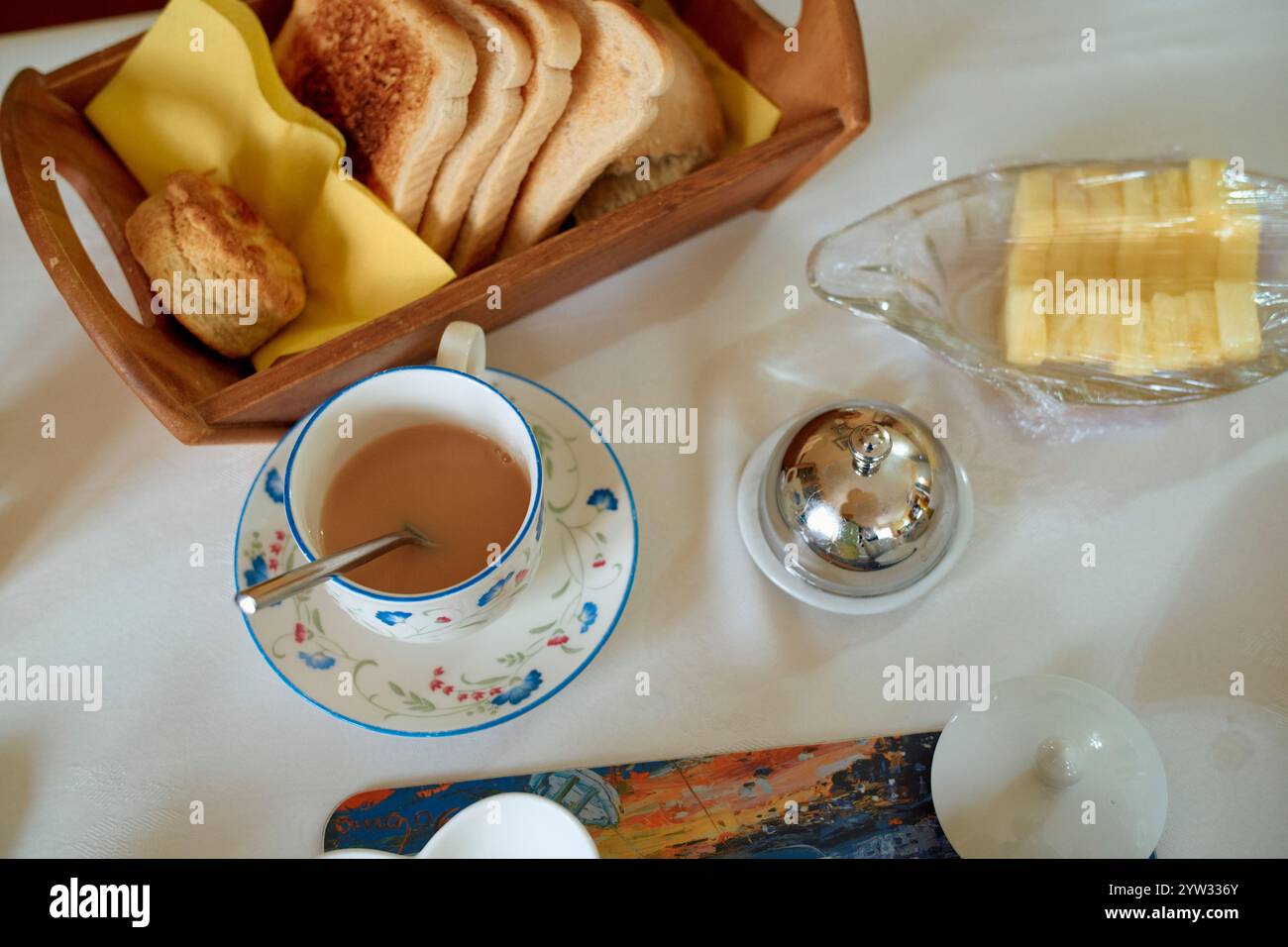 Un petit déjeuner confortable avec une tasse de thé avec du lait, du pain tranché dans un panier, du beurre et une bougie allumée sur une nappe blanche, Écosse Banque D'Images