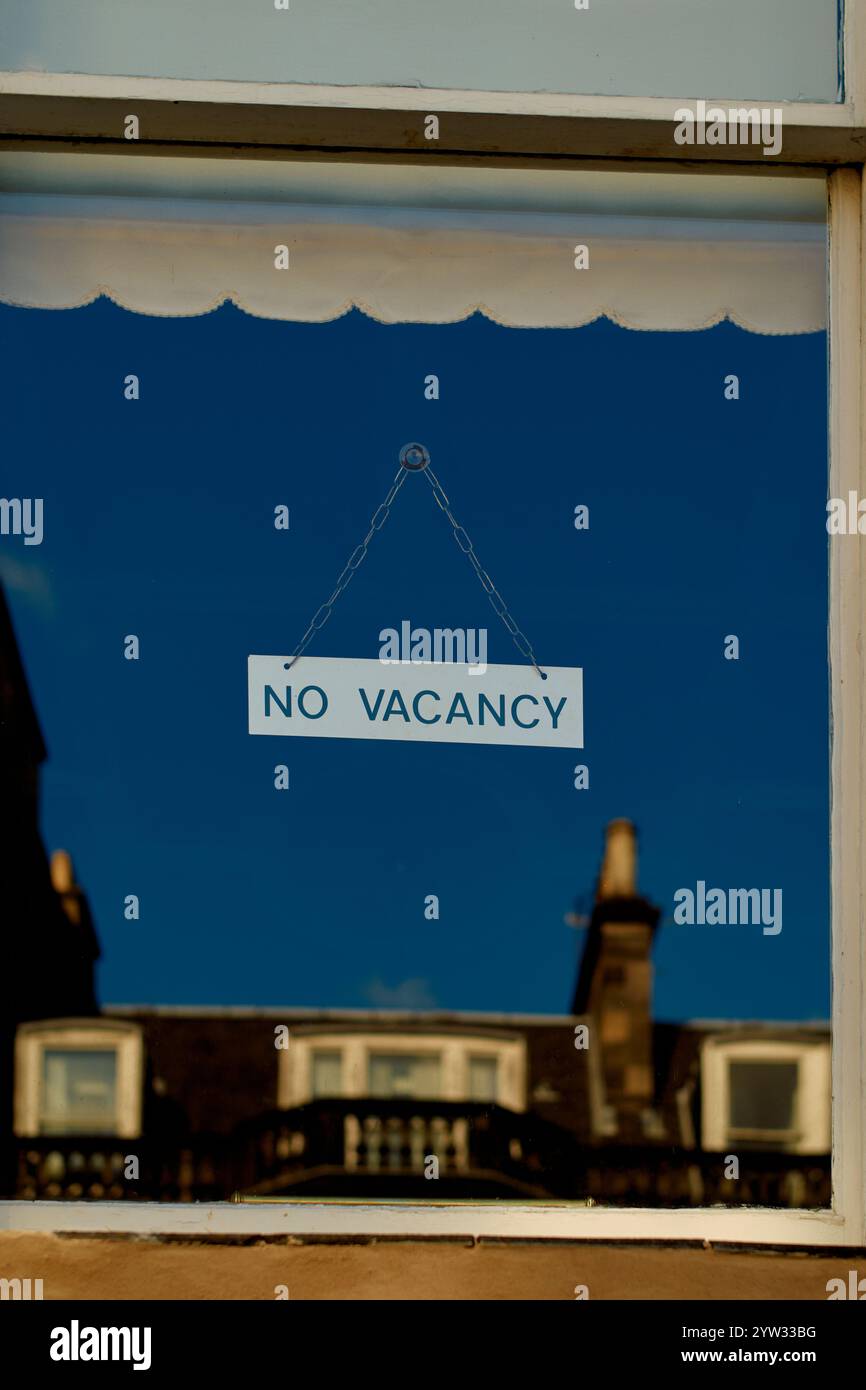 Un panneau No Vacancy est accroché à une fenêtre contre un ciel bleu clair, reflétant les bâtiments et un cadre de fenêtre rideau, Édimbourg, Écosse Banque D'Images