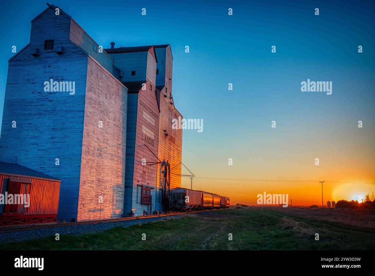 Un train descend une voie à côté d'un grand silo à grains. Le soleil se couche en arrière-plan, projetant une lueur chaude sur la scène. Le train est c Banque D'Images