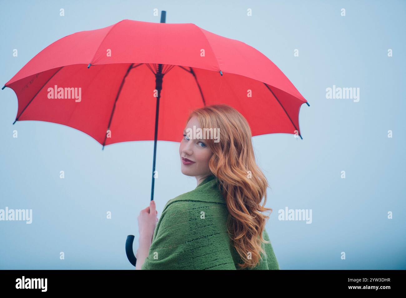 Femme aux cheveux rouges souriant par-dessus son épaule tout en tenant un parapluie rouge sur un fond bleu brumeux. Banque D'Images