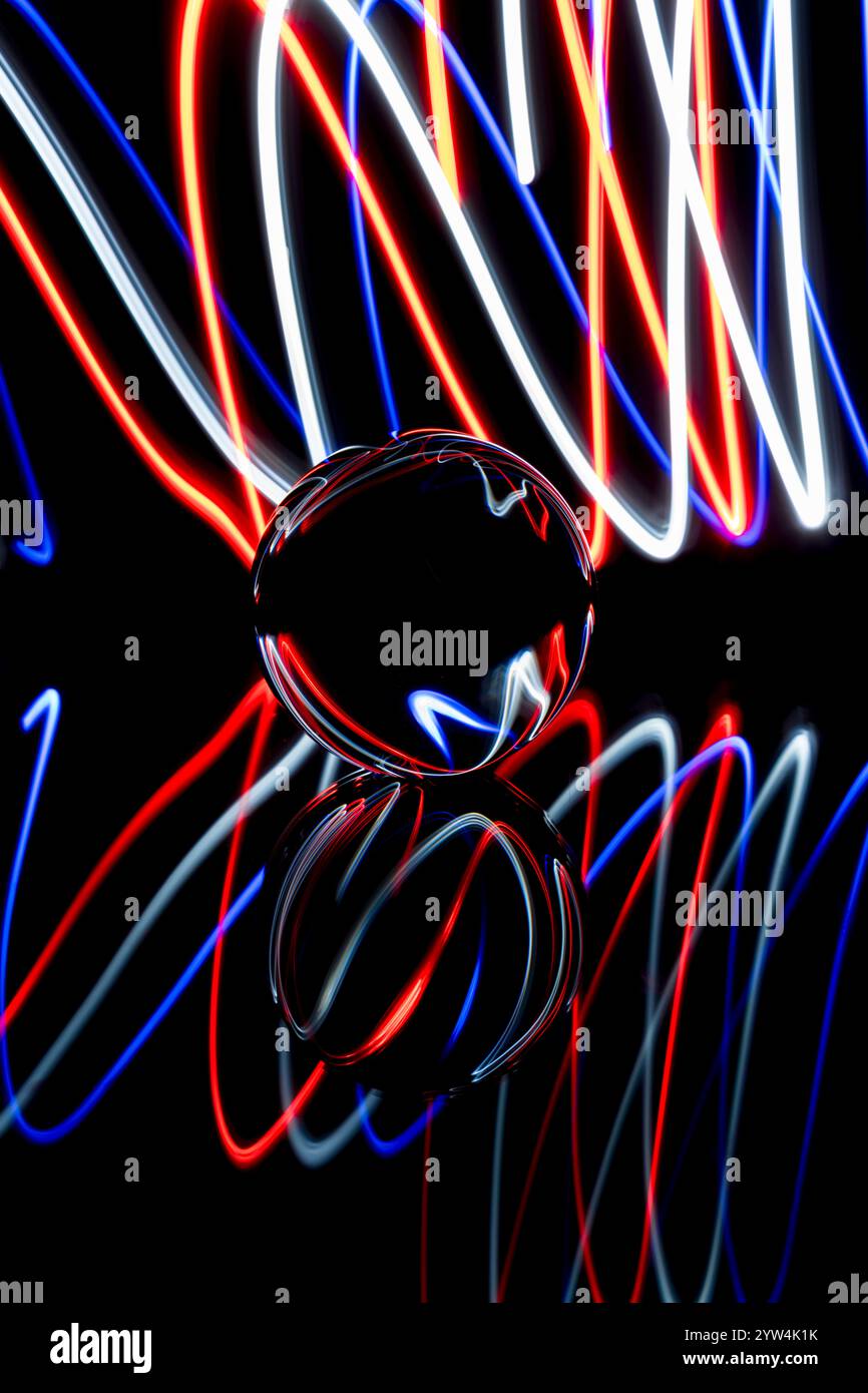 Traînées de lumière rouge, blanche et bleue abstraites créées par la peinture à la lumière autour d'une boule de verre statique posée sur un miroir sur un fond noir Uni Banque D'Images