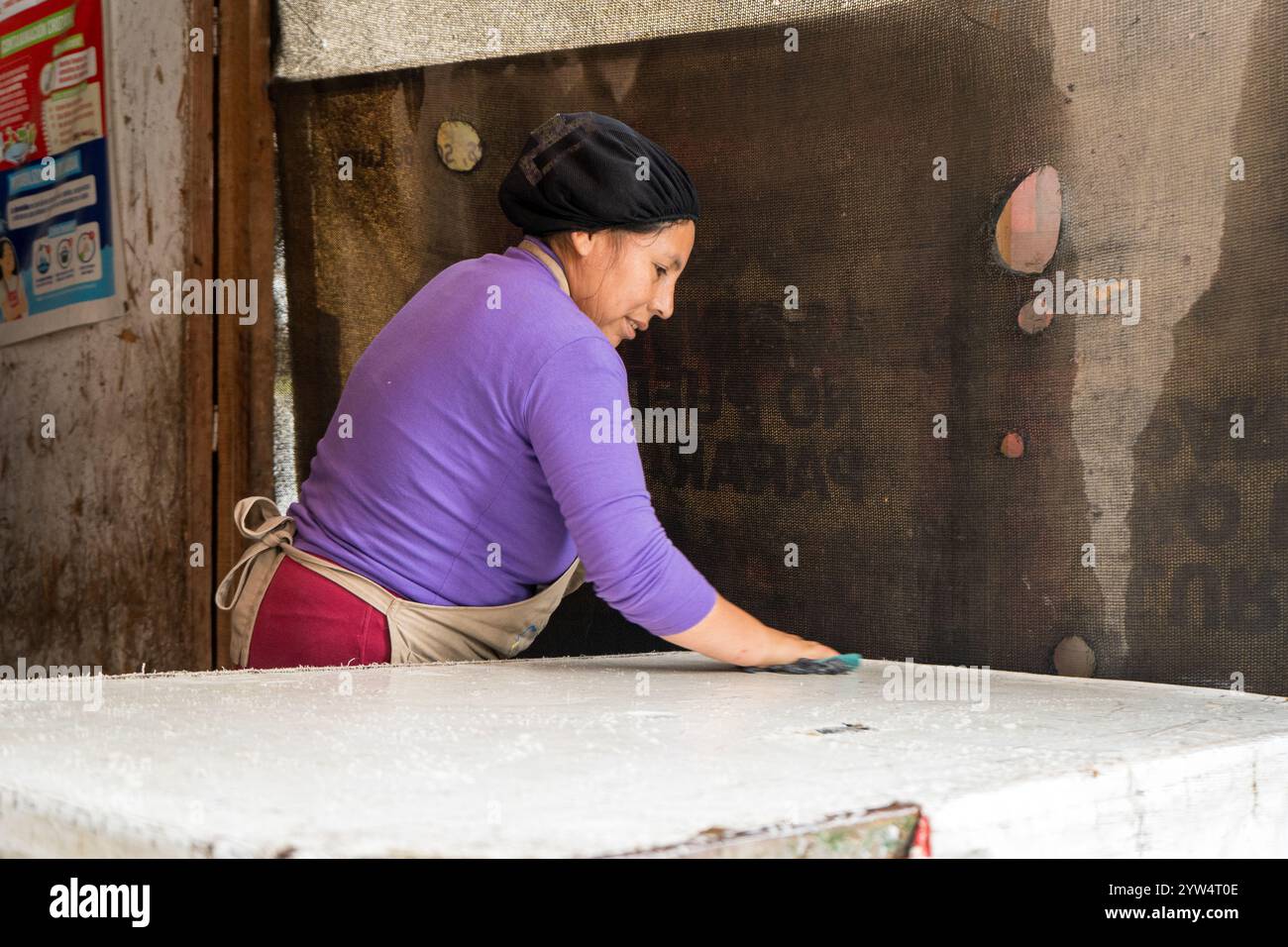 Lurin, Pérou — 6 septembre 2024 : une jeune femme en chemise violette et tablier de cuisine nettoie une table blanche dans la cuisine communautaire modeste Banque D'Images