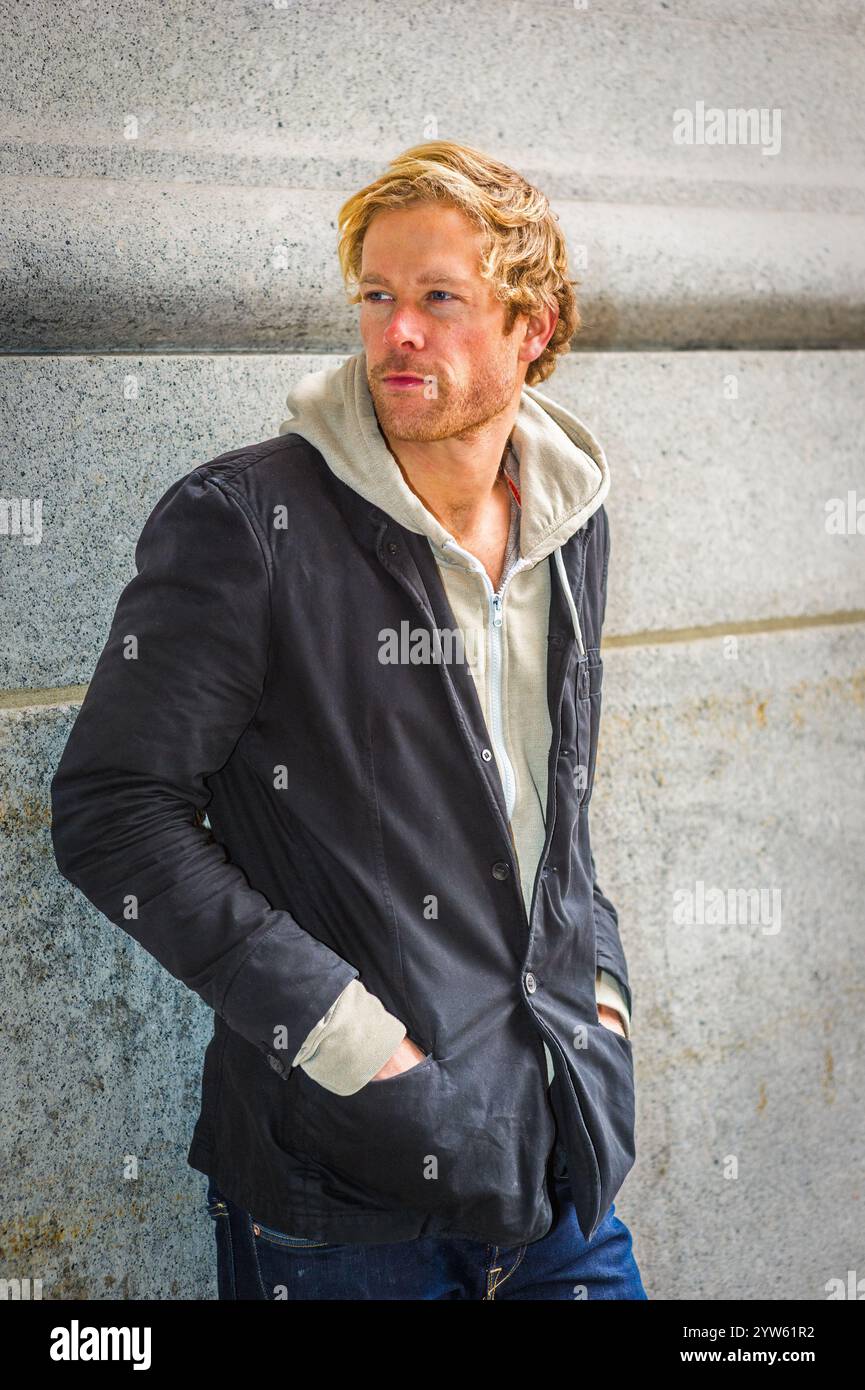 Une personne aux cheveux clairs est appuyée contre un mur de pierre, portant une veste sombre sur un sweat à capuche avec les mains dans les poches. Ils semblent regarder th Banque D'Images