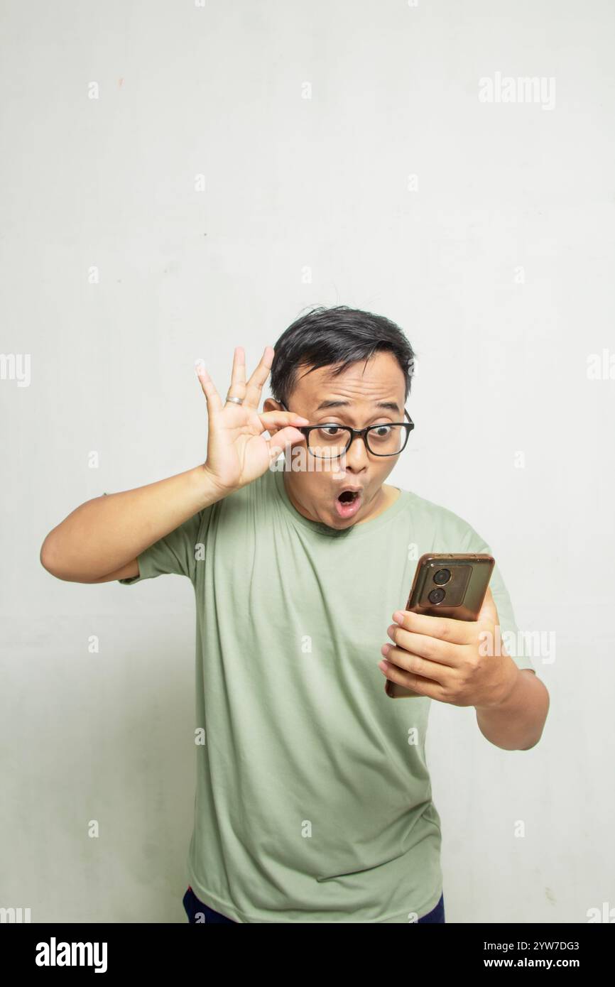 Portrait d'homme asiatique surpris avec des lunettes regardant ce qu'il voit sur son smartphone sur fond blanc de mur de studio, homme en t-shir vert clair Banque D'Images