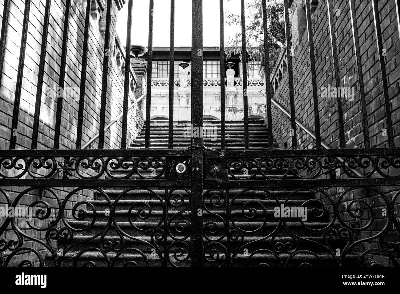 Photo en noir et blanc d'une porte en fer ornée menant à un escalier et à la façade d'un bâtiment historique, encadrée par des murs de briques. Castillo Hidalgo, Santiago. Banque D'Images