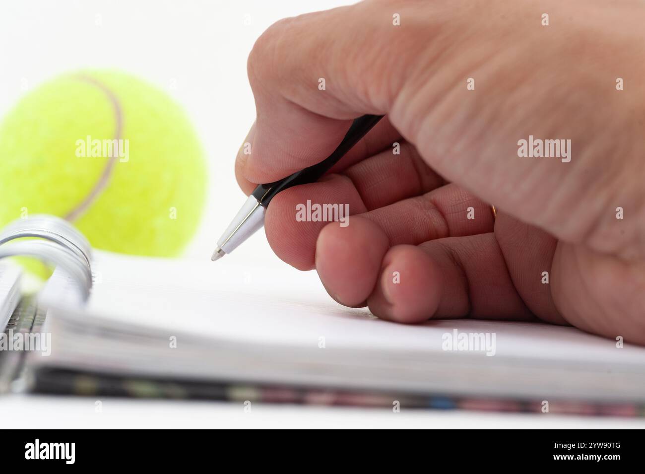 Une personne écrit dans un cahier avec un stylo. Une balle de tennis est assise sur la table à côté de l'ordinateur portable Banque D'Images