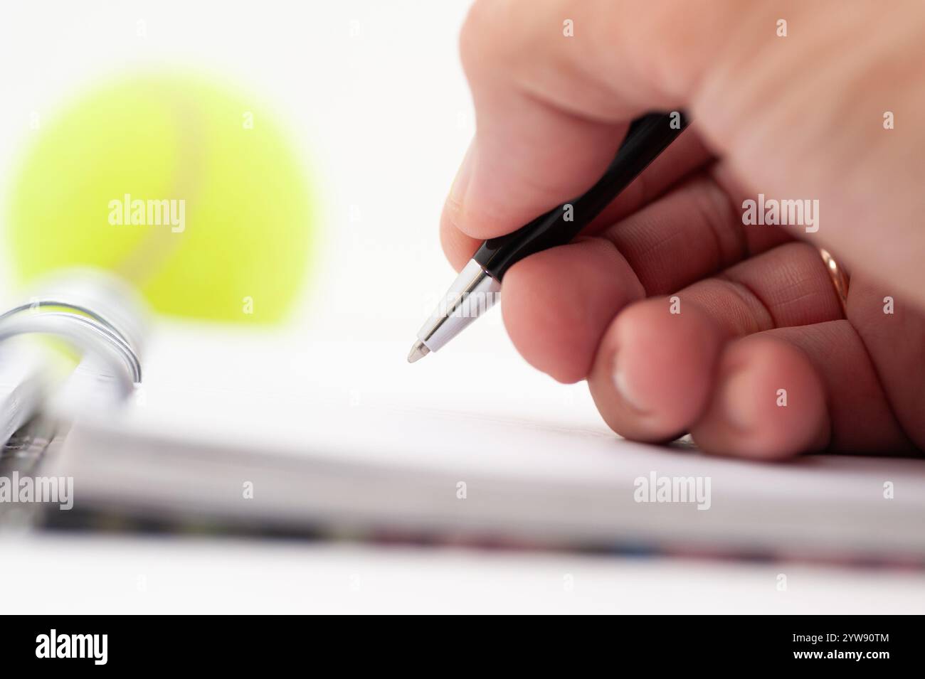 Une personne écrit dans un cahier avec un stylo. Une balle de tennis est assise sur la table à côté de l'ordinateur portable Banque D'Images