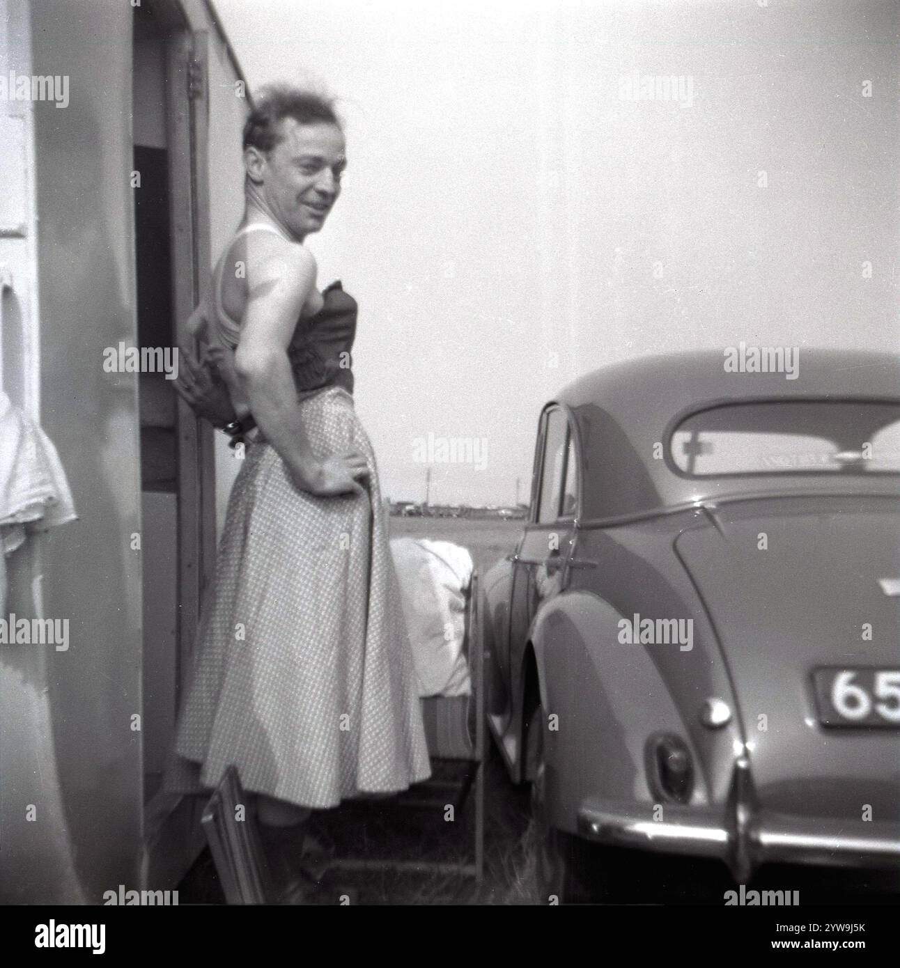 Années 1950, historique, plaisir de vacances, un homme portant un corsage féminin sur son gilet et une jupe de femme debout sur une marche devant la porte de sa caravane de vacances, une voiture de l'époque garée à côté, Angleterre, Royaume-Uni. Banque D'Images