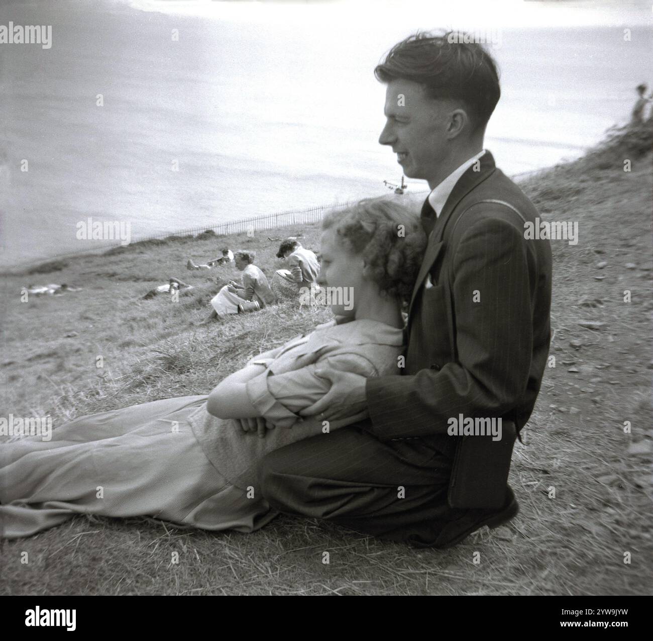 Années 1950, historique, une jeune femme dans une jupe longue et top couchée sur les genoux d'un jeune homme portant un costume à rayures épingle, chemise et cravate agenouillée sur la pente herbeuse d'une colline sur la côte, Angleterre, Royaume-Uni. Banque D'Images