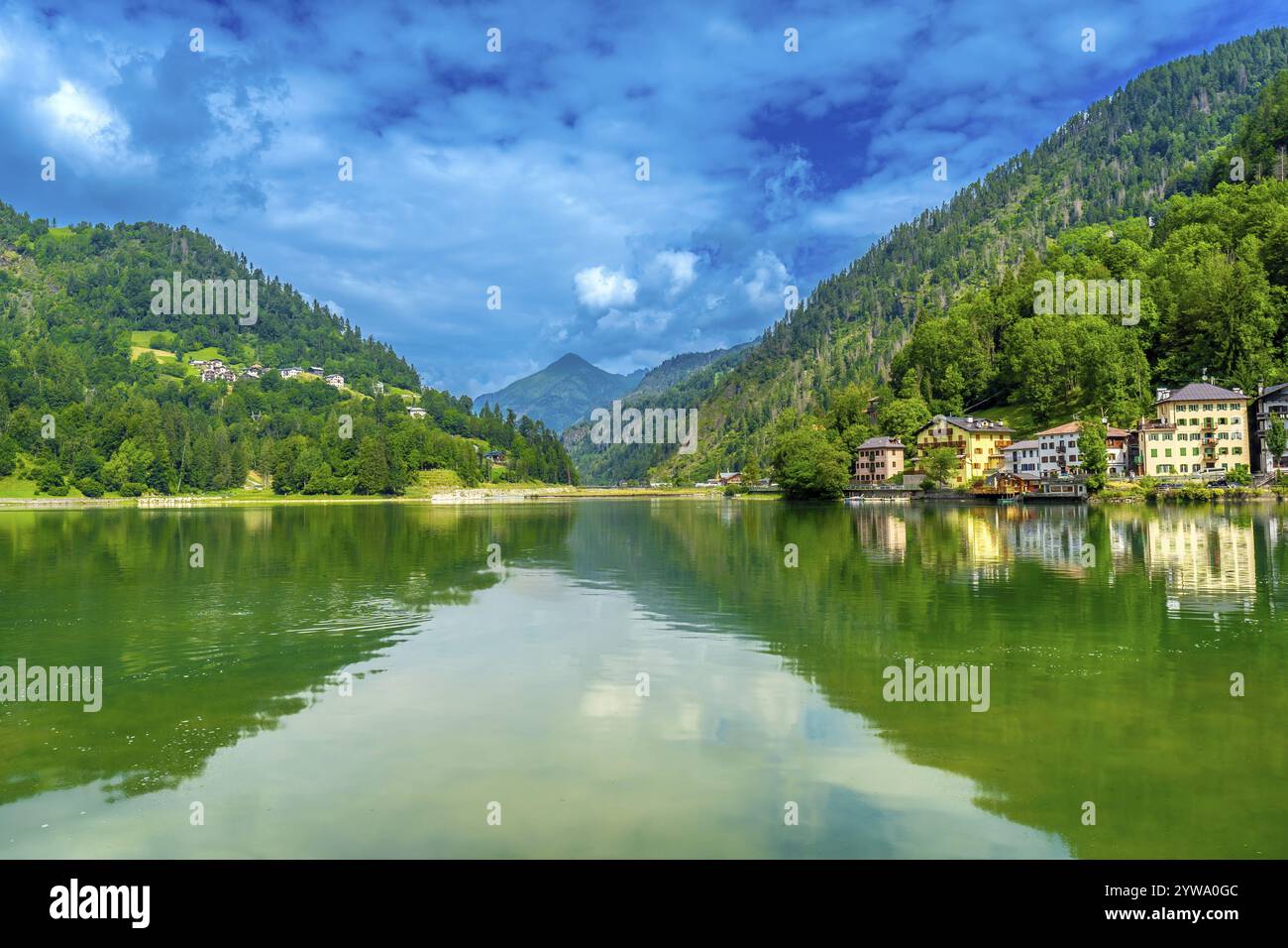 Scène estivale tranquille au lac alleghe avec des maisons colorées reflétées dans l'eau émeraude, entouré de montagnes verdoyantes sous un ciel bleu vibrant Banque D'Images