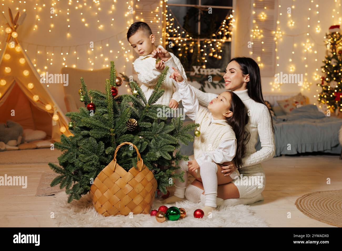 Une mère et ses deux enfants décorant un petit sapin de Noël dans une pièce chaleureuse et festive ornée de lumières de fées. Les enfants atteignent ludique Banque D'Images