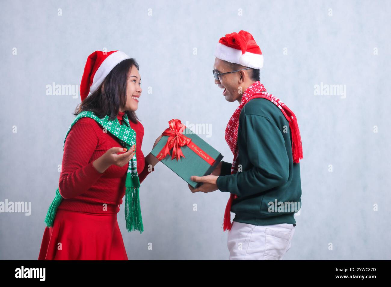 deux jeunes hommes asiatiques portant des pulls de noël, des chapeaux de père noël et des foulards, se regardant joyeusement, son ami lui donnant un cadeau de boîte verte, isolent Banque D'Images